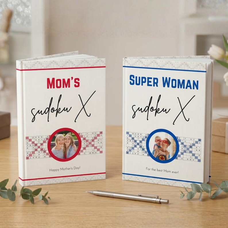 May include: Two personalized Sudoku X books with white covers. One reads "MOM'S" and "Happy Mother's Day!" with a photo. The other says "SUPER WOMAN" and "For the best Mom ever!" with a photo. A silver pen rests nearby.