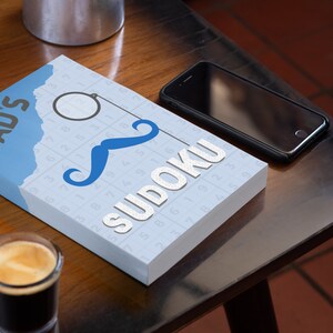 Pu&ograve; includere: Un libro blu e bianco con il testo "DAD'S SUDOKU" sulla copertina. Il libro ha un baffo blu e un monoccolo sulla copertina. Il libro &egrave; su un tavolo di legno con un bicchiere di caff&egrave; e uno smartphone nero.