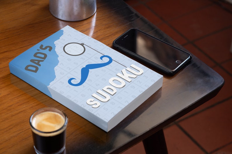 Pu&ograve; includere: Un libro blu e bianco con il testo "DAD'S SUDOKU" sulla copertina. Il libro ha un baffo blu e un monoccolo sulla copertina. Il libro &egrave; su un tavolo di legno con un bicchiere di caff&egrave; e uno smartphone nero.