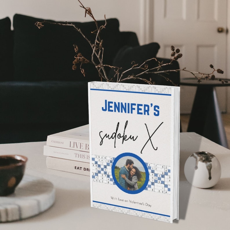 May include: A personalized sudoku book with the text "JENNIFER'S sudoku X" in blue and black. The cover features a photo of a couple inside a blue circle, sudoku puzzles, and the phrase "With love on Valentine's Day."