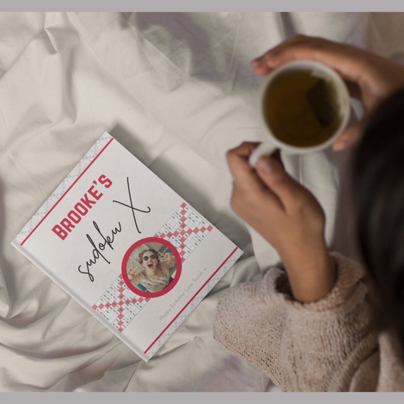 May include: A personalized Sudoku puzzle book with the text "Brooke's Sudoku X" in red and black. The cover features a photo in a red circle and a Sudoku grid design. The book is on a white surface, and a person is holding a white mug.