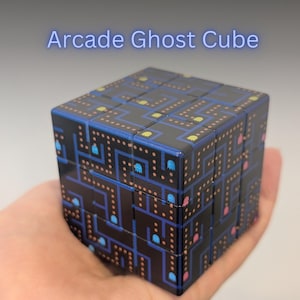 May include: A black cube puzzle with a Pac-Man-themed design. The cube features a maze pattern with blue, yellow, and pink ghosts. The text "Arcade Ghost Cube" is displayed in blue neon font.