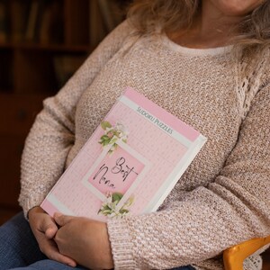 May include: A pink and white book with the text "Best Nana" and "Sudoku Puzzles" on the cover. The book is decorated with a floral design.