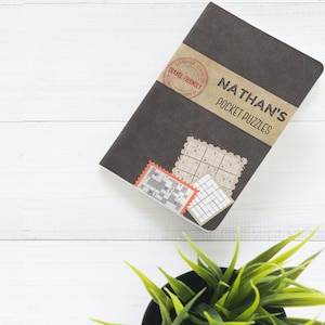 Może przedstawiać: Czarny notes z napisem "NATHAN'S POCKET PUZZLES" na okładce. Notes ma brązową papierową etykietę z czerwoną pieczątką z napisem "TRAVEL FRIENDLY".