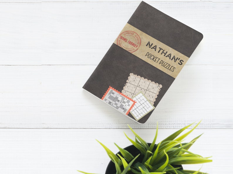 Może przedstawiać: Czarny notes z napisem "NATHAN'S POCKET PUZZLES" na okładce. Notes ma brązową papierową etykietę z czerwoną pieczątką z napisem "TRAVEL FRIENDLY".