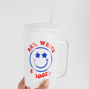 May include: White plastic cup with a red, white, and blue smiley face design. The cup has a handle and says "Red, White & Boozy" on it. It comes with a clear plastic straw.