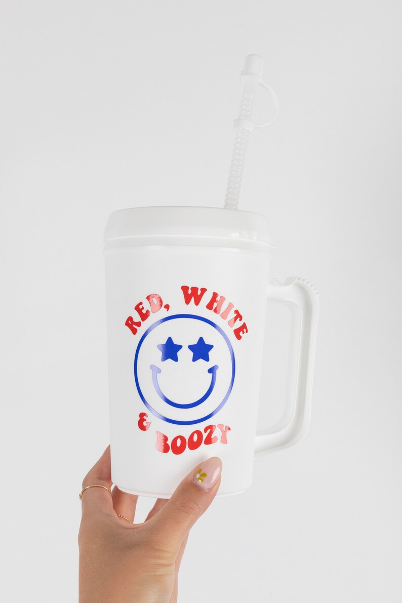 May include: White plastic cup with a red, white, and blue smiley face design. The cup has a handle and says "Red, White & Boozy" on it. It comes with a clear plastic straw.