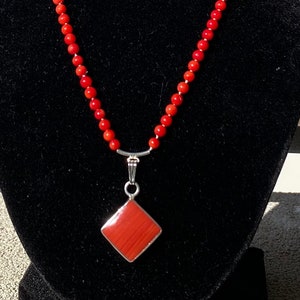 May include: A vibrant red beaded necklace with a diamond-shaped pendant. The pendant is a reddish-orange color, set in a silver-toned metal. The necklace is displayed on a black velvet bust against a sunny background.