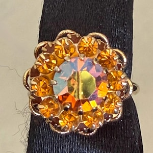 May include: A gold-toned ring featuring a large, central iridescent stone surrounded by a halo of smaller, round, amber-coloured faceted stones. The setting has a scalloped, floral-inspired edge that frames the circular arrangement of crystals.