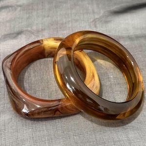 May include: Two bangle bracelets made of translucent amber-colored resin. One bracelet features a smooth, rounded circular shape, while the other has a slightly squared, geometric profile. Both pieces exhibit a swirling, marbled pattern throughout.