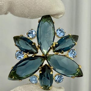 Op de afbeelding: Een bloemvormige broche met grote, marquise-geslepen blauwgroene kristallen in een sterpatroon, geaccentueerd door kleinere, ronde, lichtblauwe kristallen in een goudkleurig metalen frame. Het ontwerp heeft een symmetrische uitstraling.