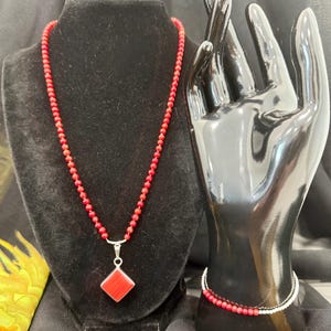 May include: A red beaded necklace with a silver pendant featuring a red square gemstone. A matching red beaded bracelet is displayed on a black hand mannequin. The jewelry is set against a black backdrop.