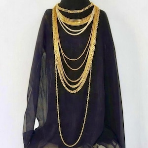 May include: An array of gold necklaces, featuring diverse chain styles and lengths, draped over a black top. The necklaces include fine chains and thicker links, creating a striking visual effect. The black fabric provides a contrasting backdrop to the gold jewellery.
