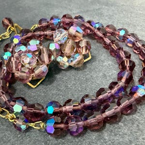 May include: A vintage jewelry set featuring a necklace and matching clip-on earrings. The necklace and earrings are made of faceted, purple beads with iridescent highlights. The necklace has a gold-tone chain and clasp.