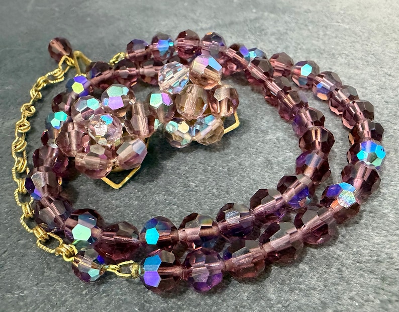May include: A vintage jewelry set featuring a necklace and matching clip-on earrings. The necklace and earrings are made of faceted, purple beads with iridescent highlights. The necklace has a gold-tone chain and clasp.