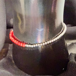 May include: A two-strand bracelet featuring red and silver beads. The red beads are a vibrant color, while the silver beads have a metallic sheen. The bracelet is displayed on a black mannequin.