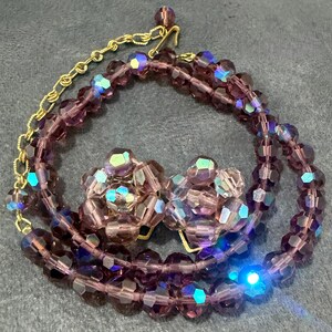 May include: A vintage jewelry set featuring a necklace and matching clip-on earrings. The necklace and earrings are made of faceted, purple beads with iridescent highlights. The necklace has a gold-tone chain.