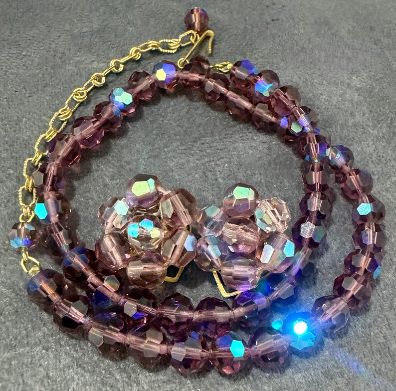 May include: A vintage jewelry set featuring a necklace and matching clip-on earrings. The necklace and earrings are made of faceted, purple beads with iridescent highlights. The necklace has a gold-tone chain.