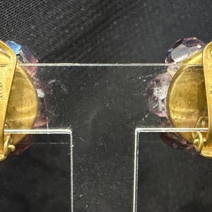 May include: A pair of vintage clip-on earrings with a cluster of faceted, lavender-colored crystals. The gold-toned metal clips are stamped "MADE ITALY". The earrings are displayed on a clear plastic stand against a dark background.