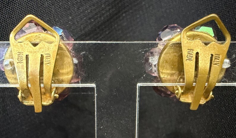 May include: A pair of vintage clip-on earrings with a cluster of faceted, lavender-colored crystals. The gold-toned metal clips are stamped "MADE ITALY". The earrings are displayed on a clear plastic stand against a dark background.