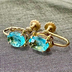 May include: A pair of gold-tone screw-back earrings featuring oval-cut, bright blue faceted stones. Each earring is set in a decorative metal frame with a threaded screw mechanism at the back for securing the jewelry to the earlobe.