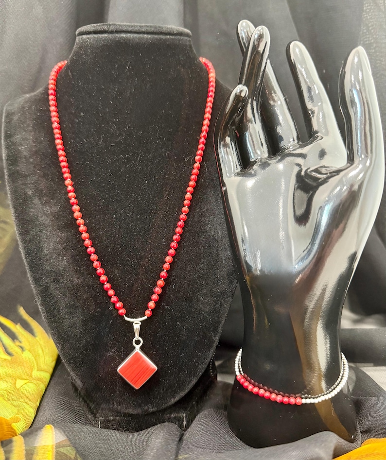 May include: A red beaded necklace with a silver pendant featuring a red square gemstone. A matching red beaded bracelet is displayed on a black hand mannequin. The jewelry is set against a black backdrop.