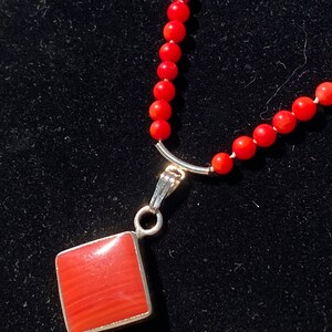 May include: A red coral bead necklace with a silver pendant. The pendant is a square shape with a red coral center and a silver border. The necklace is on a black background.