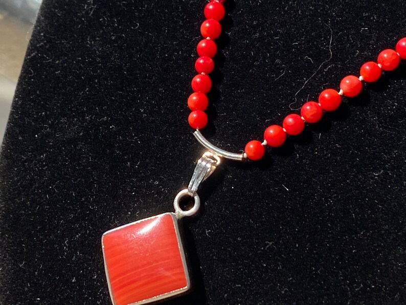 May include: A red coral bead necklace with a silver pendant. The pendant is a square shape with a red coral center and a silver border. The necklace is on a black background.
