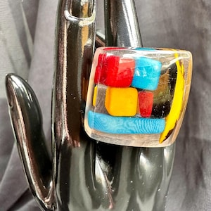 May include: A clear resin ring with colorful geometric shapes. The ring features red, blue, and yellow blocks encased in clear resin. The ring is displayed on a black hand.