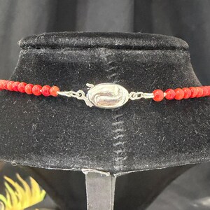 May include: A red beaded necklace with a silver clasp. The necklace is displayed on a black velvet bust. The beads are small and round, and the clasp is a decorative oval shape.