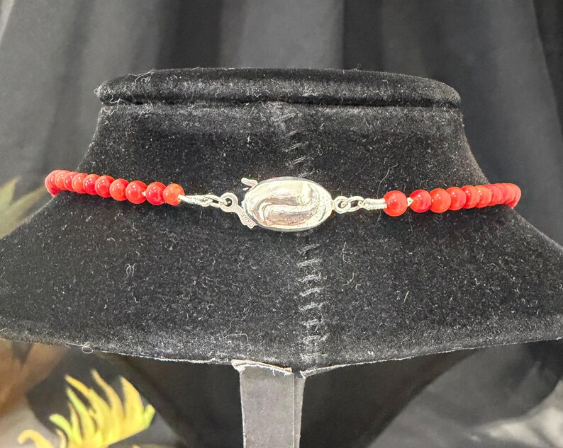 May include: A red beaded necklace with a silver clasp. The necklace is displayed on a black velvet bust. The beads are small and round, and the clasp is a decorative oval shape.