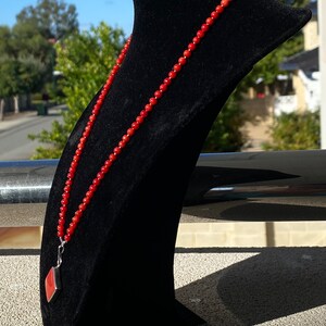 May include: A red beaded necklace with a silver pendant featuring a red stone, displayed on a black velvet bust. The necklace is composed of small, round beads. The pendant has a geometric shape.