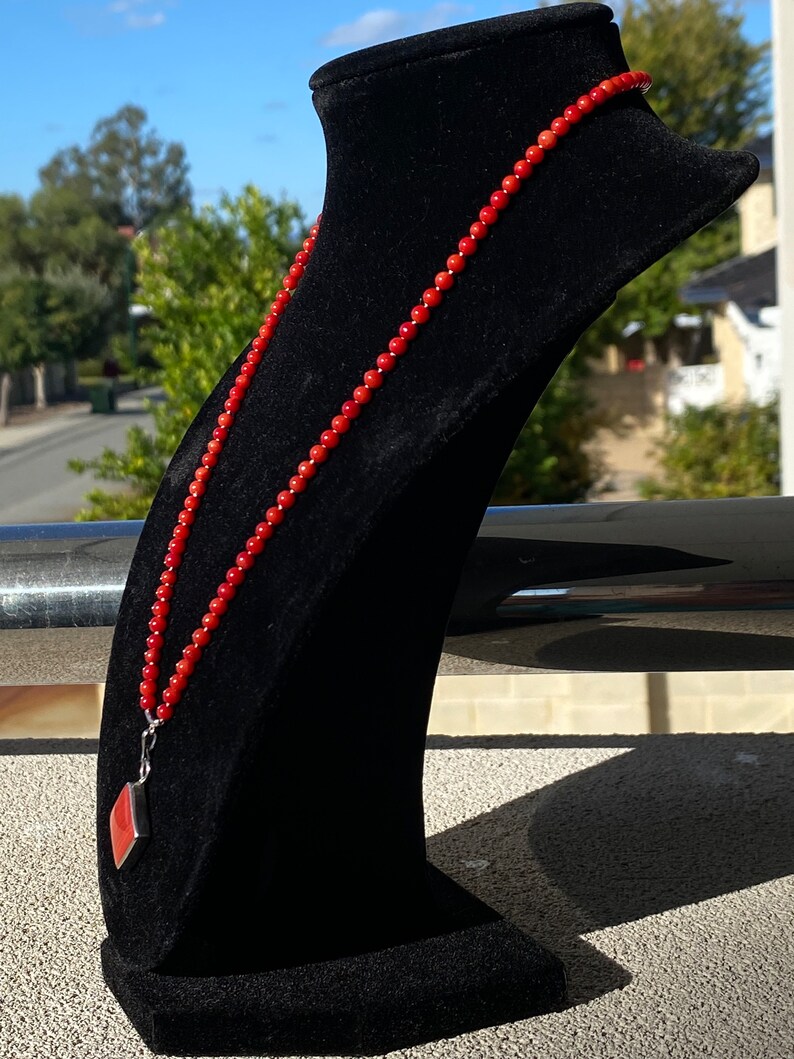 May include: A red beaded necklace with a silver pendant featuring a red stone, displayed on a black velvet bust. The necklace is composed of small, round beads. The pendant has a geometric shape.