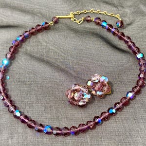 May include: A vintage jewelry set featuring a purple beaded necklace and matching clip-on earrings. The necklace has a gold-tone clasp. The beads have an iridescent quality, reflecting blue and purple hues. The earrings are clustered with similar beads.