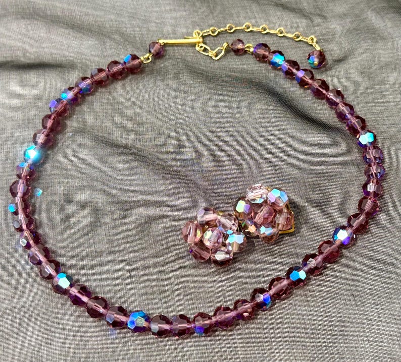 May include: A vintage jewelry set featuring a purple beaded necklace and matching clip-on earrings. The necklace has a gold-tone clasp. The beads have an iridescent quality, reflecting blue and purple hues. The earrings are clustered with similar beads.