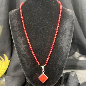 May include: A red beaded necklace with a silver pendant. The pendant is a red square set in a silver frame. The necklace is displayed on a black velvet bust.
