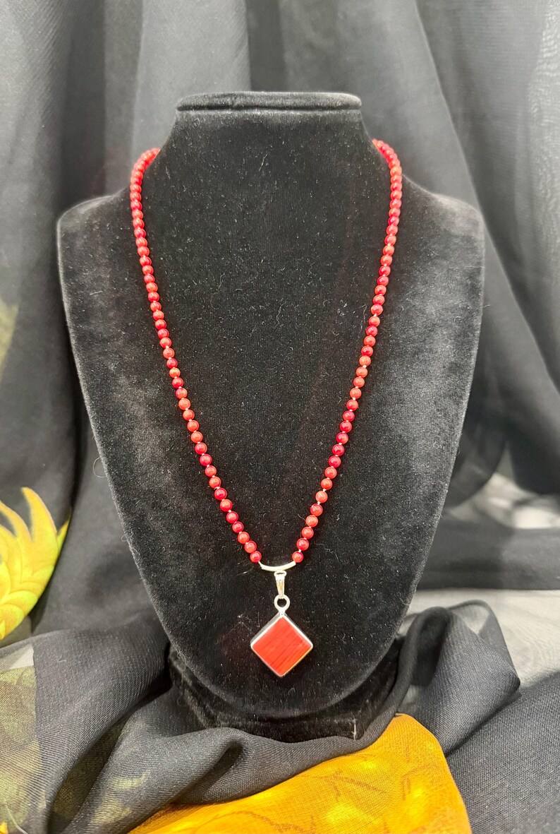 May include: A red beaded necklace with a silver pendant. The pendant is a red square set in a silver frame. The necklace is displayed on a black velvet bust.