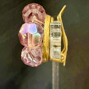 May include: A clip-on earring featuring three faceted pink beads and a gold-tone metal clip. The beads have an iridescent quality, reflecting multiple colors. The earring is designed with a gold-tone metal band and a clear, rectangular clip.