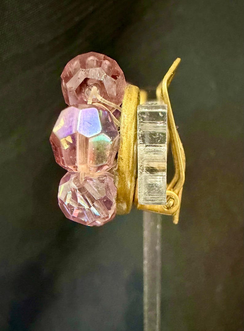 May include: A clip-on earring featuring three faceted pink beads and a gold-tone metal clip. The beads have an iridescent quality, reflecting multiple colors. The earring is designed with a gold-tone metal band and a clear, rectangular clip.