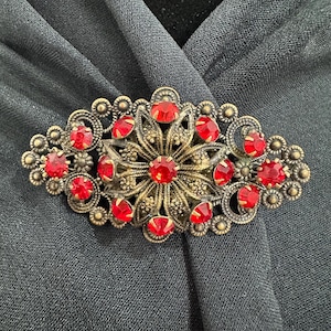 Op de afbeelding: Vintage broche met een bloemenontwerp. De broche heeft een goudkleurige metalen basis versierd met talrijke ovaalvormige, helderrode edelstenen. De broche is ovaal van vorm en is geplaatst tegen een donkergrijze stoffen achtergrond.