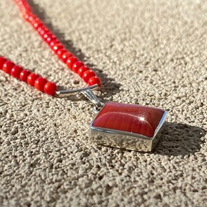 May include: A red beaded necklace with a rectangular pendant. The pendant has a red, polished stone set in a silver-colored metal frame. The necklace is composed of small, round red beads.