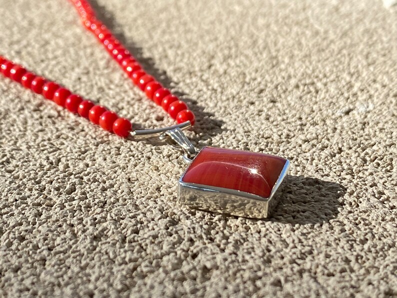 May include: A red beaded necklace with a rectangular pendant. The pendant has a red, polished stone set in a silver-colored metal frame. The necklace is composed of small, round red beads.