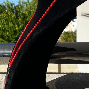 May include: A vibrant red beaded necklace displayed on a black velvet jewelry stand. The necklace is composed of small, round beads and hangs gracefully. The background features greenery and a bright sky, highlighting the necklace's color.