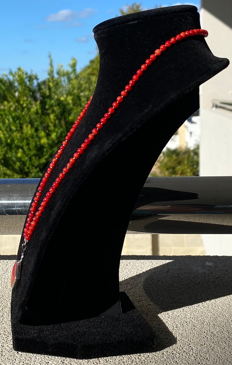May include: A vibrant red beaded necklace displayed on a black velvet jewelry stand. The necklace is composed of small, round beads and hangs gracefully. The background features greenery and a bright sky, highlighting the necklace's color.