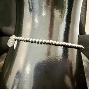 May include: A silver beaded bracelet with a small, round tag that reads "925". The bracelet is made of small, round beads strung together. The bracelet is displayed on a black surface.