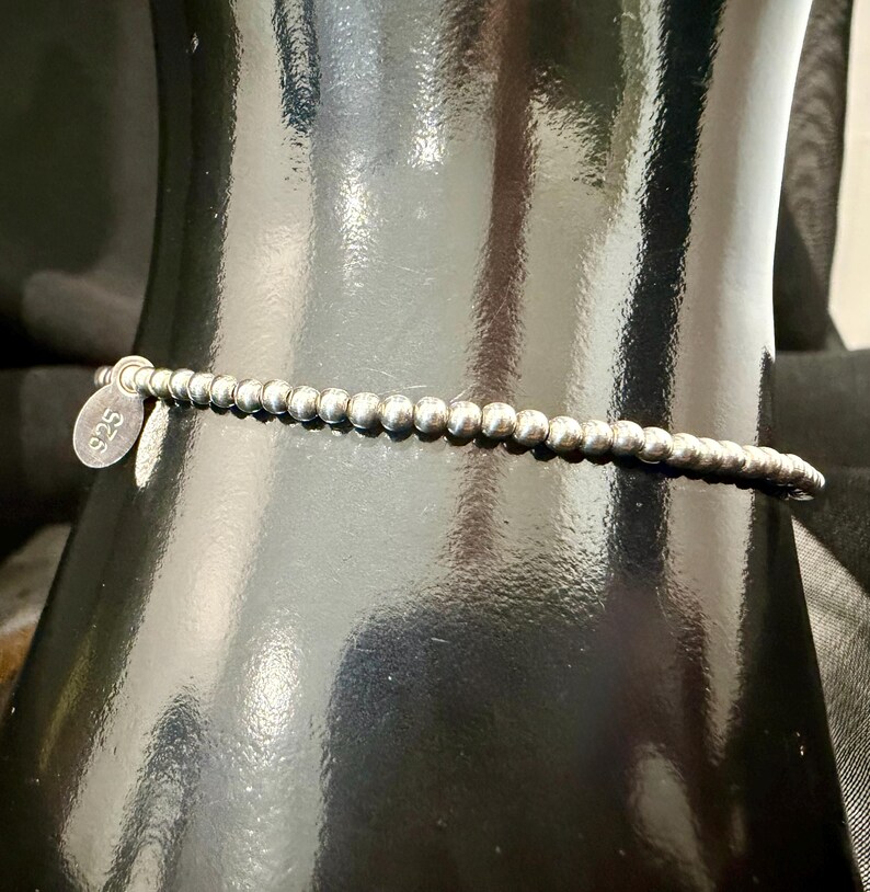May include: A silver beaded bracelet with a small, round tag that reads "925". The bracelet is made of small, round beads strung together. The bracelet is displayed on a black surface.