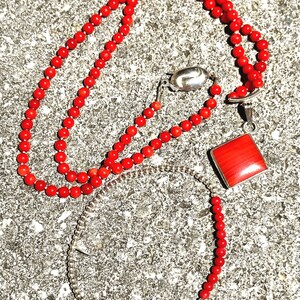 May include: A red coral bead necklace, bracelet, and pendant set. The necklace and bracelet are made of small, round red beads. The pendant is a square of red coral set in a silver frame. The jewelry is displayed on a textured gray surface.