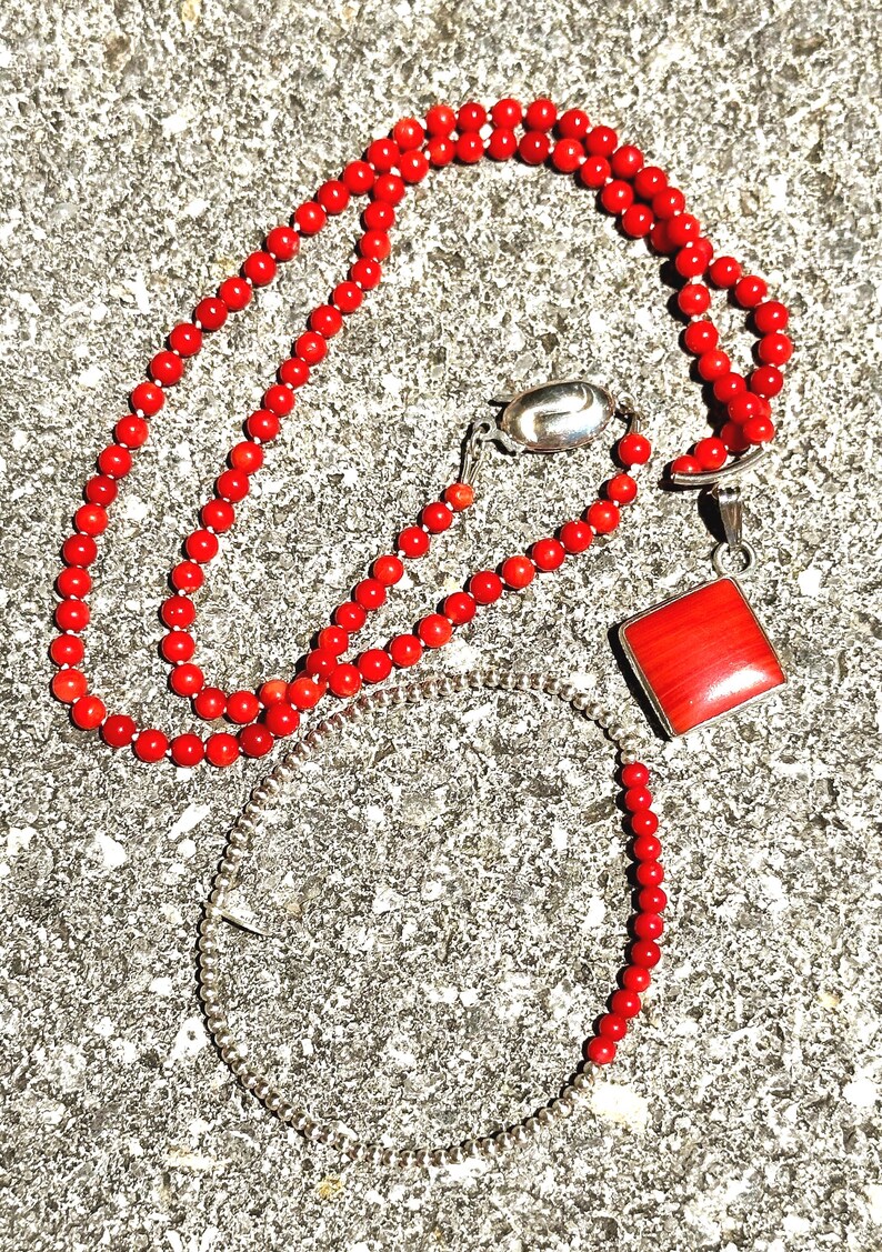 May include: A red coral bead necklace, bracelet, and pendant set. The necklace and bracelet are made of small, round red beads. The pendant is a square of red coral set in a silver frame. The jewelry is displayed on a textured gray surface.