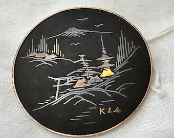 Op de afbeelding: Een zwart rond sieraad met een zilveren en gouden ontwerp van een Japans berglandschap met een pagode en een torii-poort. Het sieraad is gemarkeerd met de letters "K24".