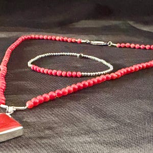 May include: A red beaded necklace with a square pendant and a matching bracelet. The pendant is a square of red material set in a silver frame. The necklace and bracelet are made of small, round red beads. The jewelry is displayed on a dark background.